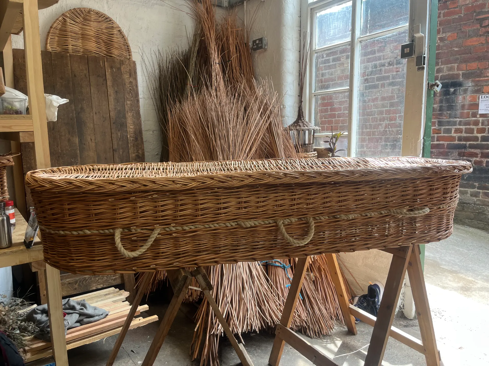 A handwoven willow coffin on trestles in the workshop, with bundles of willow behind